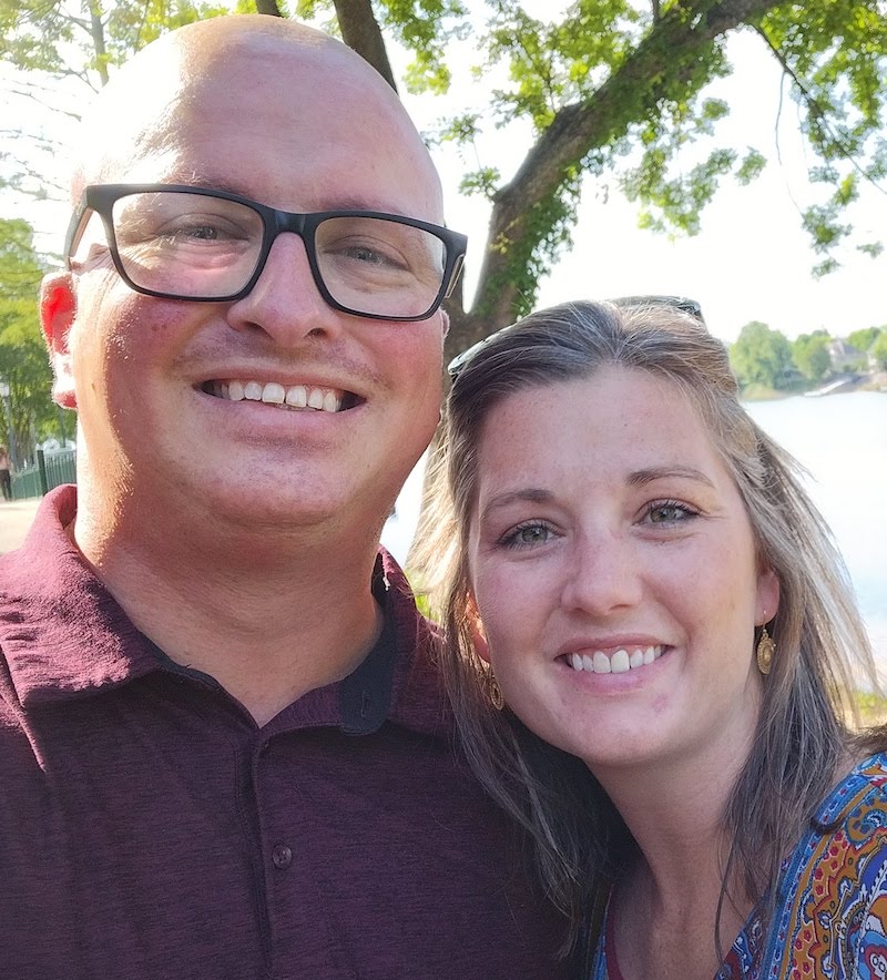 two folks smiling at the camear. one is bald and bespectacled, and the other, a female, is wearing sunglasses holding back her long hair. a lake is blurrily visible behind them.