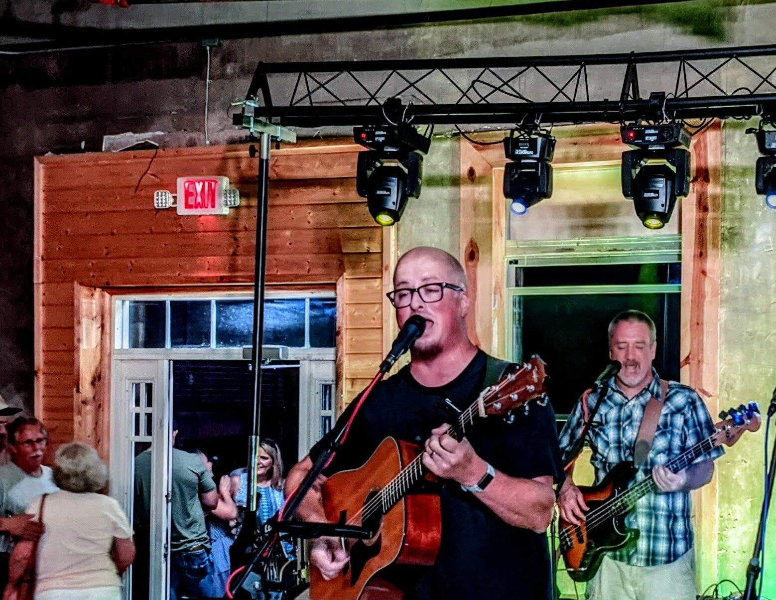 a bald man plays guitar and sings into a microphone on stage, complete with stage lighting above him. 